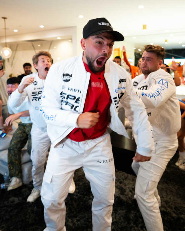 A group of excited men in matching white outfits celebrate energetically in a brightly lit room, with one man in a black cap at the center shouting joyfully.